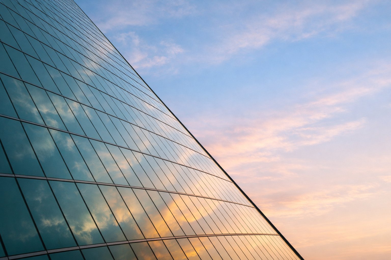 Luminous glass facade angled against open sky, sunlight catching architectural planes with visible blue and amber sky above